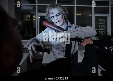 Spooky and aggressive man with zombie greasepaint on his face and hands fighting with angry businessman holding baseball bat Stock Photo