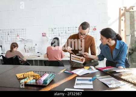 Young contemporary fashion designers choosing colors and textile for new collection against two females using sewing machines in workshop Stock Photo