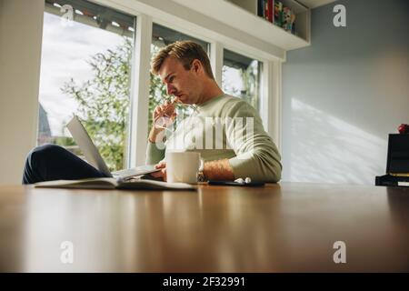Pensive young man sitting at table and looking at his laptop. Caucasian male using laptop and thinking. Stock Photo