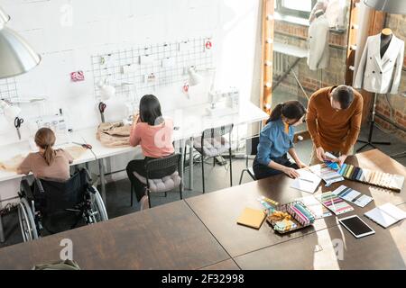 Two young contemporary fashion designers choosing colors for new collection while their female colleagues using sewing machines Stock Photo