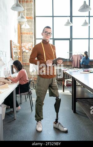 Young fashion designer or tailor with prosthetic leg using tablet by workplace against female colleagues inside large workshop Stock Photo