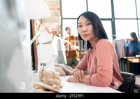 Young Asian seamstress in casualwear sitting on chair by workplace inside large contemporary workshop and using sewing machine Stock Photo