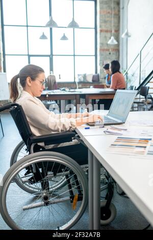 Young creative designer in wheelchair using laptop while drawing sketches for new seasonal collection against two colleagues in workshop Stock Photo