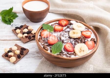 Chocolate cornflakes with milk and strawberry in wooden bowl on white wooden background and linen textile. Side view, close up. Stock Photo