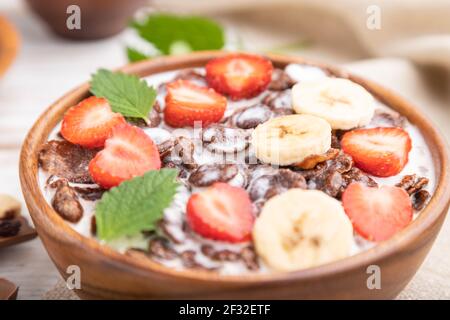 Chocolate cornflakes with milk and strawberry in wooden bowl on white wooden background and linen textile. Side view, close up, selective focus. Stock Photo