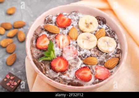 Chocolate cornflakes with milk, strawberry and almonds in ceramic bowl on gray concrete background and orange linen textile. Side view, close up, sele Stock Photo