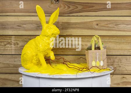 yellow Easter bunny on white semicircular wooden table with feathers, twigs and a bag with plants. Wooden planks as a background. Stock Photo