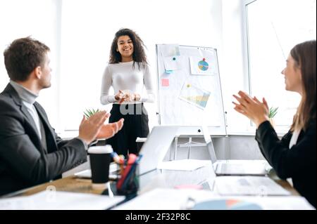 Happy successful african american woman is standing near flipchart on briefing meeting, smiling. Beautiful stylish young business lady making a presentation or submitting work report on conference Stock Photo