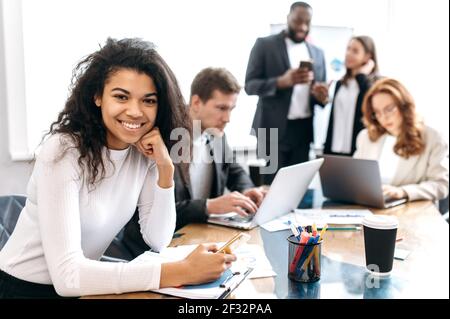 Portrait of young elegant business woman at the workplace. Beautiful female employee is sitting at the work desk in office at briefing meeting, hold a smartphone in hand, smiling Stock Photo