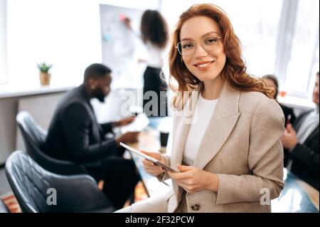 Young adult stylish business woman is standing in office at briefing, using tablet, searching ideas. Beautiful redhead lady working on corporate project with colleagues, wearing elegant suit Stock Photo