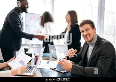 Happy businessman in formal suit is sitting in office at the conference, learning financial graphs. Young adult male manager is looking directly at the camera and smile Stock Photo
