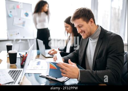 Joyful male employee doing paperwork on business meeting, smiling. Successful young manager in formal suit learning financial graphs, working with collegues on corporate project Stock Photo