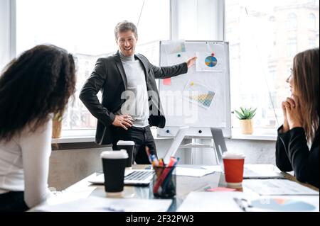Caucasian young adult businessman is making presentation on conference, talking about financial graphs. Confident male employee in stylish suit submit work report on briefing meeting Stock Photo