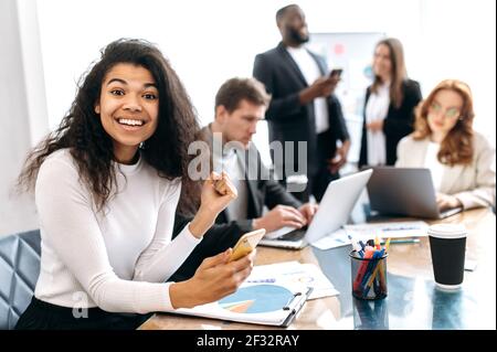 Excited young african american lady using smartphone, sitting in office. Joyful beautiful businesswoman got job promotion, done successful project, look at the camera, smiling Stock Photo