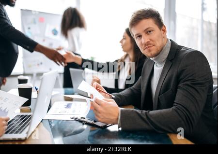 Young caucasian businessman on briefing meeting, sitting at the work desk, is learning financial graphs. Handsome male employee looking at the camera, smiling, doing paperwork Stock Photo