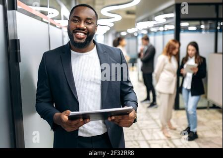 Handsome successful businessman in formal stylish suit is standing in office hall, smiling. Attractive african american male employee looking at the camera, hold tablet in arms Stock Photo