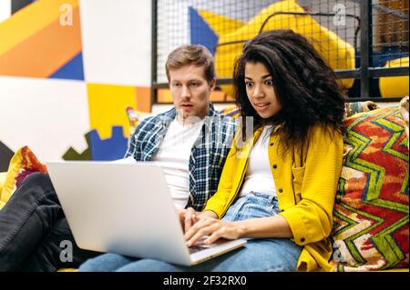 Shocked male and female employees sitting on the sofa, looking at the laptop screen. Multiethnic business people got unexpected message or browsing internet and reading surprised news Stock Photo