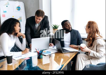 Influential business team in modern office on briefing, working on new project. Successful male and female employees discussing ideas, learning financial graphs, using laptop Stock Photo