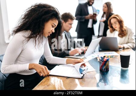 Concentrated african american woman doing paperwork, sitting in modern office on conference. Focused business lady learning financial graphs, working on corporate project at briefing meeting Stock Photo