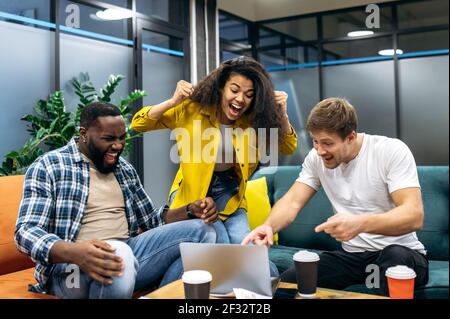 Excited young adult business people sitting in office, looking at the laptop screen, feeling euphoric. Overjoyed employees or students passing important exams or get big profit from corporate project Stock Photo