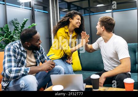 Satisfied multiracial students or colleagues sitting in modern office, well done with corporate project. Successful coworkers giving high five to each other, smiling, happy about good work Stock Photo
