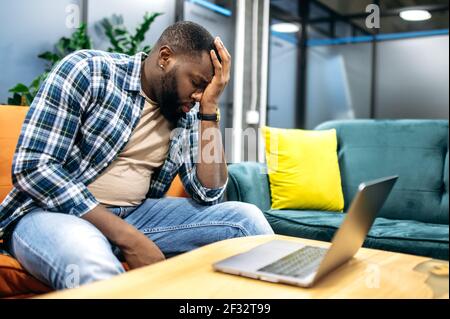 Tired african american employee or student is sitting on the sofa, having a headache. Exhausted freelancer needing a rest, working on tense project, overwork concept Stock Photo