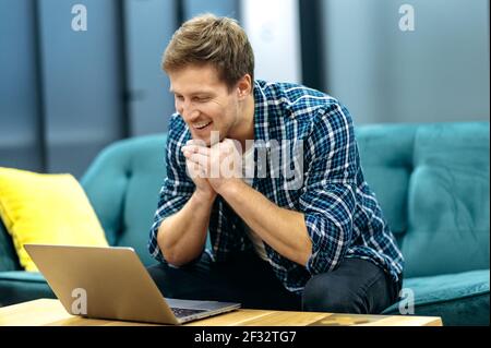 Satisfied male freelancer is using laptop, having online meeting. Stylish caucasian businessman is sitting on the sofa, smiling, talking with colleagues, working on corporate project Stock Photo