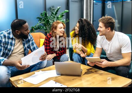Multiracial young adult colleagues sitting on the sofa, discussing work questions. Happy friendly male and female employees having a conversation, talking about corporate project, teamwork concept Stock Photo