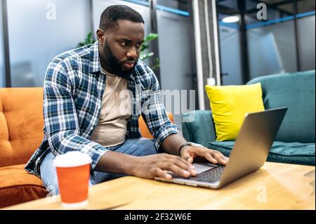 Serious african american freelancer is using laptop, working on important project. Concentrated male employee sitting in modern office, developing new app or business plan for company Stock Photo