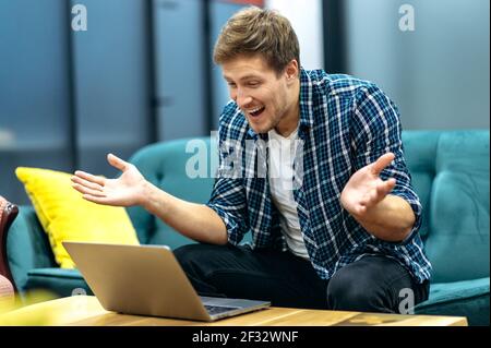 Caucasian male employee is sitting on the sofa, using laptop. Smiling young businessman are having online conference, emotionally discussing project with colleagues, communication concept Stock Photo