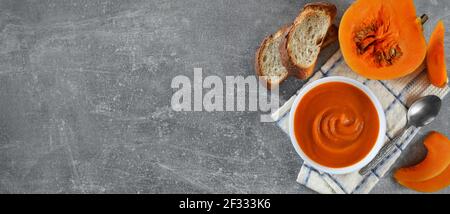 Vegetarian pumpkin soup in a white bowl next to fresh pumpkin and slices of bread on a grey background with copy space, flat lay. Healthy eating conce Stock Photo