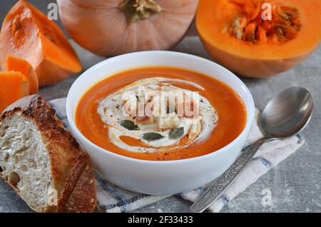 Appetizing pumpkin cream soup with croutons, sour cream and pumpkin seeds in a white bowl next to fresh pumpkin and a slice of bread. Healthy eating c Stock Photo