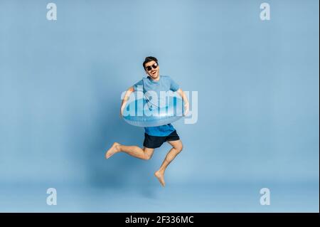 Full length photo of a joyful happy caucasian guy in a sunglasses, a blue t-shirt and with a blue inflatable ring, jumping high on a isolated blue background, travel, beach vacation concept Stock Photo