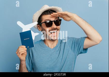 Cheerful happy caucasian young man in sunglasses and a summer hat, holds a passport and a small plane in his hand, looks out in anticipation of a long-awaited summer vacation, isolated blue background Stock Photo