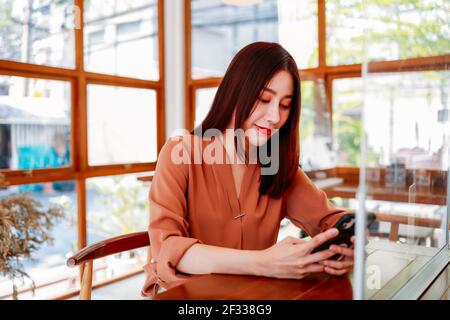 Beautiful young Asian woman sitting in modern cafe for a break while relaxing and browsing smartphone with mirror between two table with social distancing due to covid-19 Stock Photo