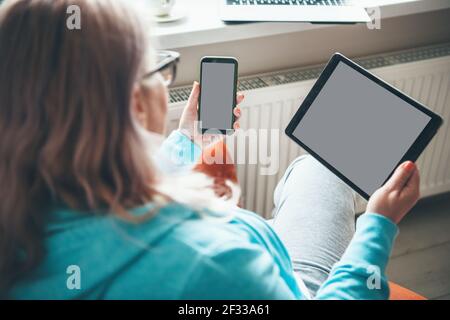 Back view photo of a caucasian senior woman working on a tablet and holding a phone at home Stock Photo
