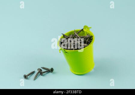 Screws and a small green bucket on a blue background. Black metal hardware in a bucket. Close-up, selective focus. Stock Photo