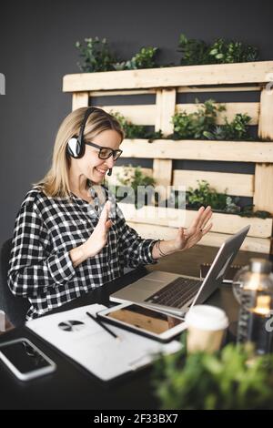 pretty, young blond businesswoman with black and white checked shirt is sitting in a sustainable, ecological office with a headset and is having a web Stock Photo