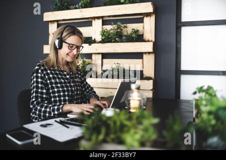 pretty, young blond businesswoman with black and white checked shirt is sitting in a sustainable, ecological office with a headset and is having a web Stock Photo