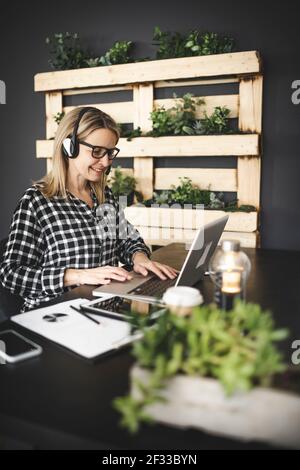 pretty, young blond businesswoman with black and white checked shirt is sitting in a sustainable, ecological office with a headset and is having a web Stock Photo