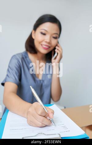 Smiling pretty spa salon or clinic reception worker talking on phone with client and filling questionnaire with his personal data of client Stock Photo