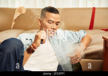 Handsome young Asian man smoking cigarette and drinking alcohol at home Stock Photo