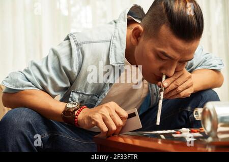 Handsome young Vietnamese man sniffing cocaine through rolled banknote at home Stock Photo