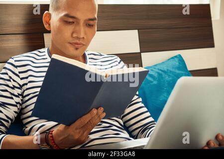 Asian student sitting on bed, working on laptop and reading interesting book Stock Photo