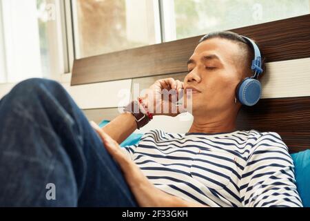 Calm young Vietnamese man relaxing on bed and enjoying good music in headphones Stock Photo