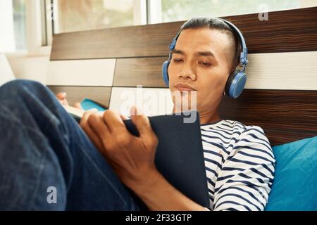 Serious young Asian man in headphones lying on bed, listening to music and reading book Stock Photo