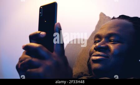 Young african american man browsing social media on his smartphone with a smile while relaxing on the couch. High quality photo Stock Photo
