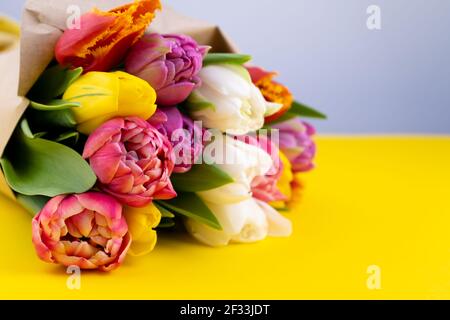 A bouquet of tulips wrapped in craft paper. Spring flowers. Gift delivery. Celebration. Copy space. Yellow, white, pink, purple buds. Yellow background. Postcard. Tulip flower Stock Photo