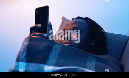 Young black man checking social media on his phone covered with blanket on the couch. High quality photo Stock Photo