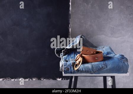 Denim jeans jacket suit and belt at shelf near grey wall background texture. Stack of classic jeans fashion at table Stock Photo